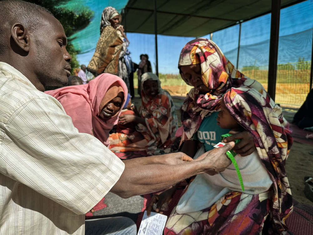 Dziecko podczas pomiaru obwodu ramienia za pomocą taśmy MUAC. Wynik w czerwonym obszarze wskazuje na niedożywienie. Sudan, 2025.